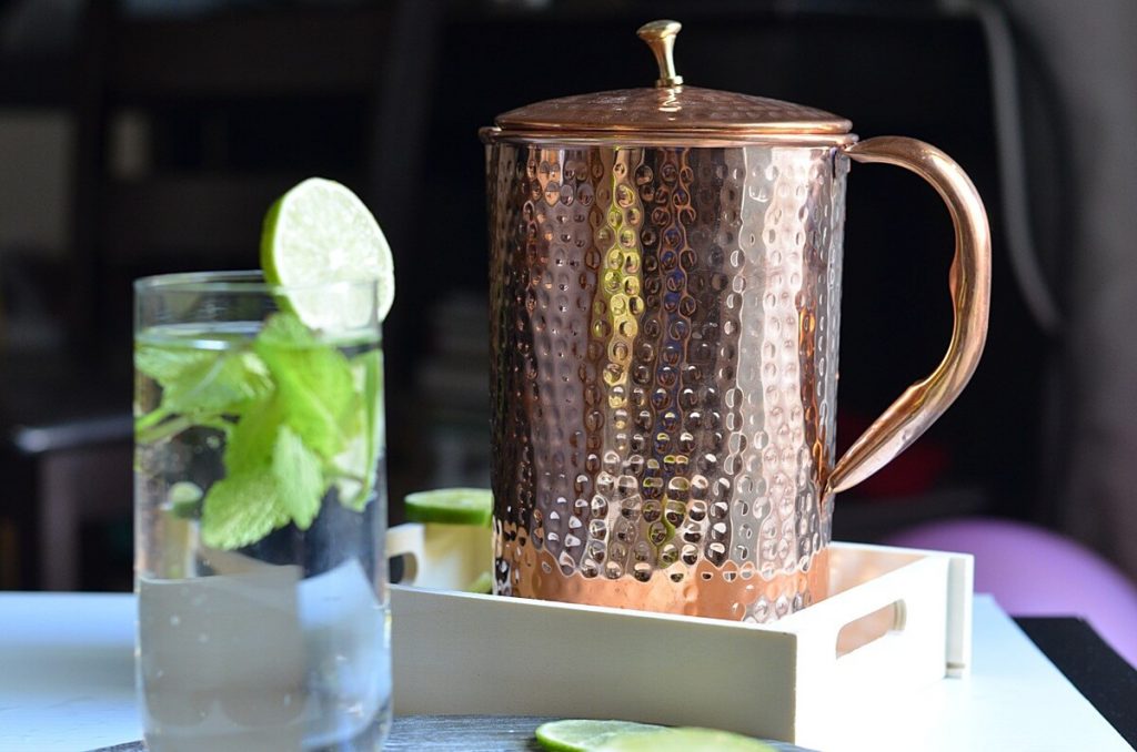 Copper pitcher is kept in a wooden tray. glass is filled with water with lemon wedges & mint leaves.