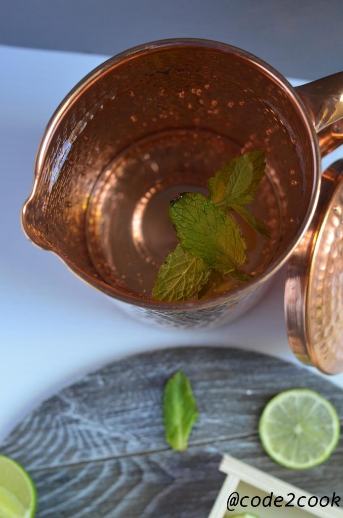 Water in the copper pitcher.Lemon wedges and mint leaves are scattered on wooden surface.