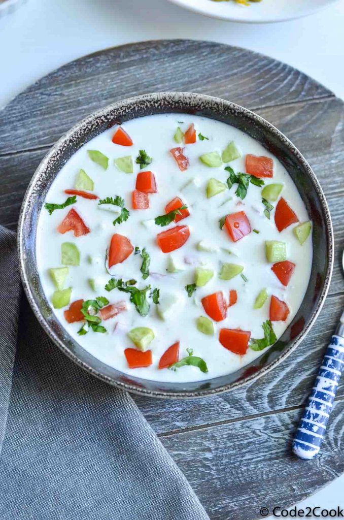 Raita is served in grey bowl, garnished with chopped cucumber,tomato & coriander.