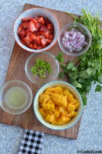 all ingredients of mango strawberry salsa are kept over wooden board.