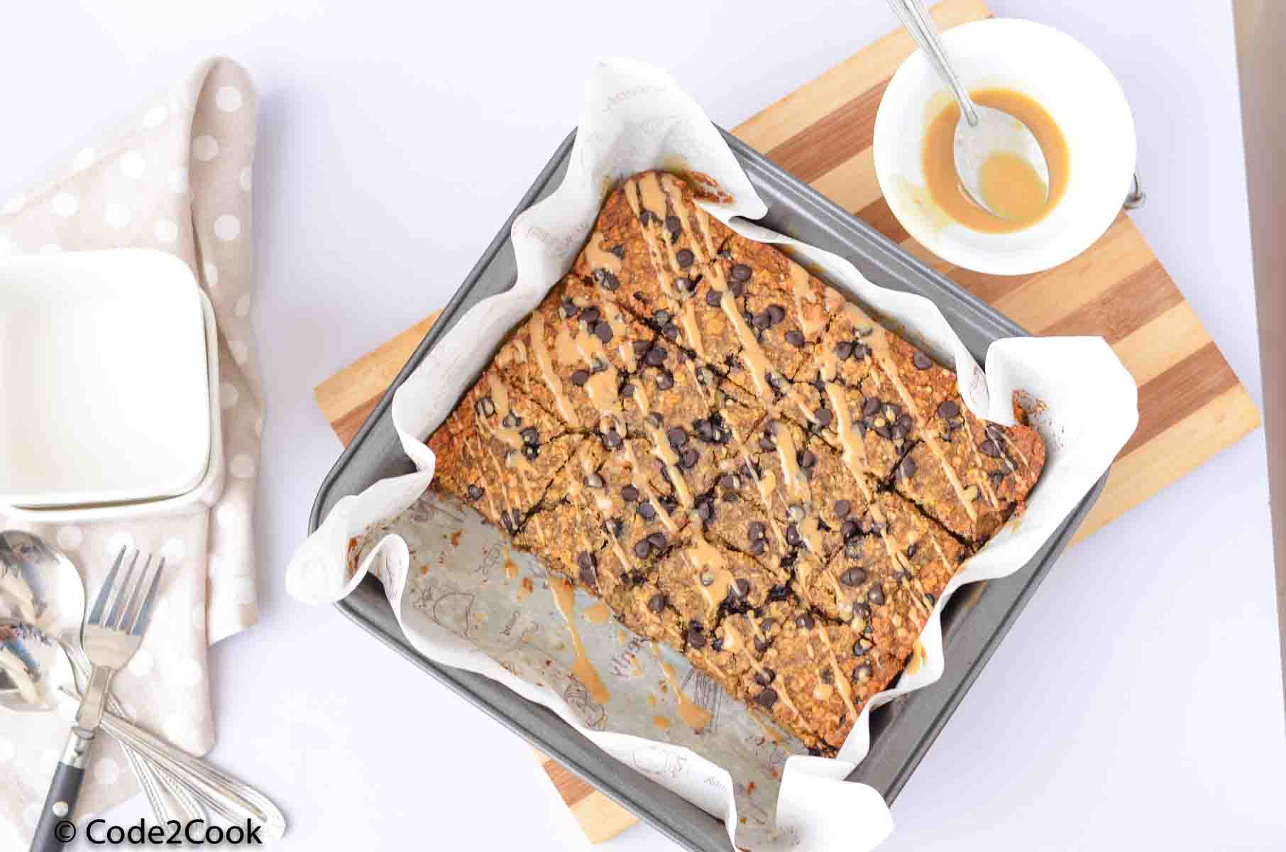 healthy oatmeal bars are in the baking pan. Peanut butter glaze is kept in a bowl and at left side.